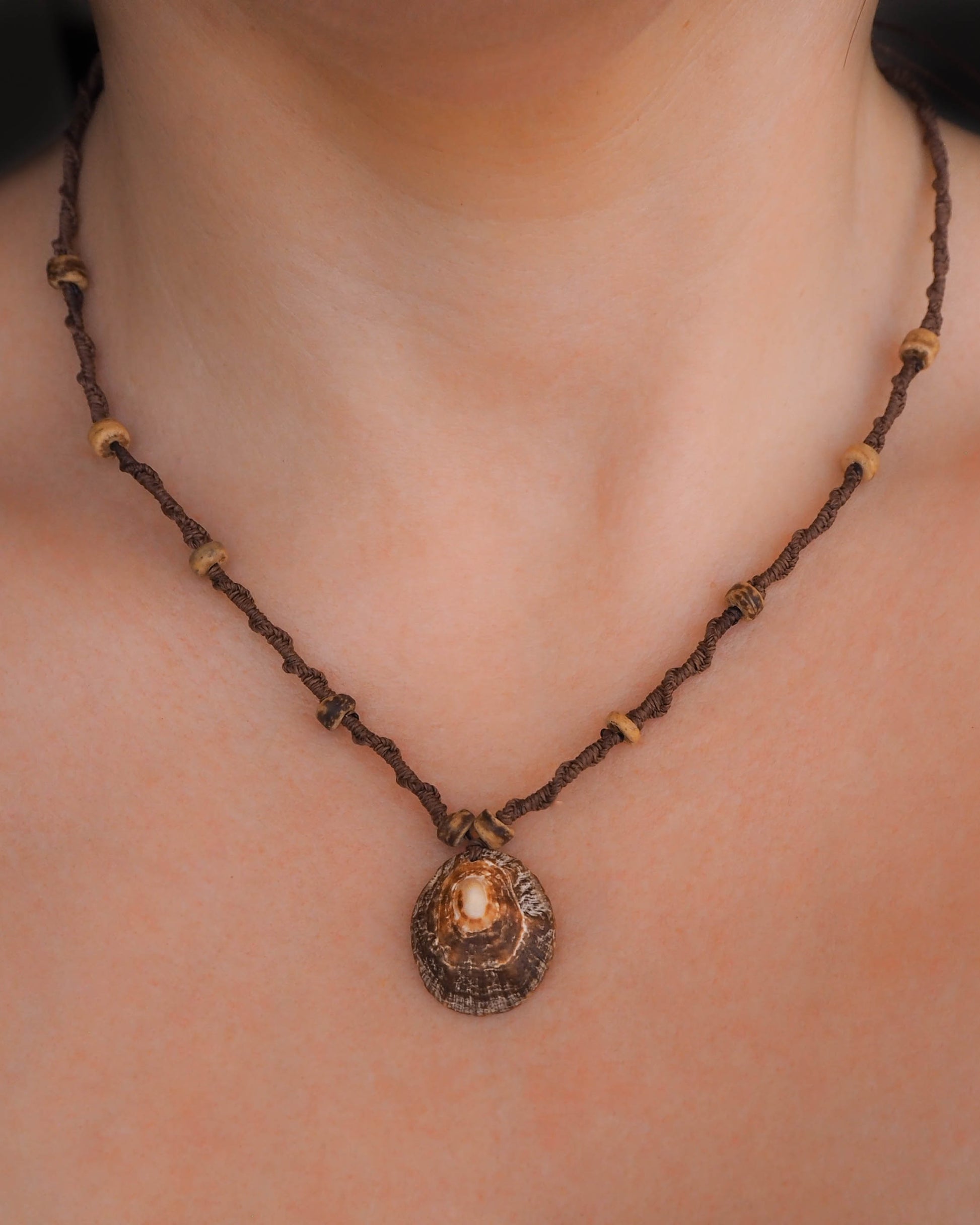close-up photo of the necklace's centerpiece: a shimmering limpet shell pendant, delicately suspended from a braided brown wax cord. Wooden beads are interspersed along the cord, adding a touch of rustic charm.